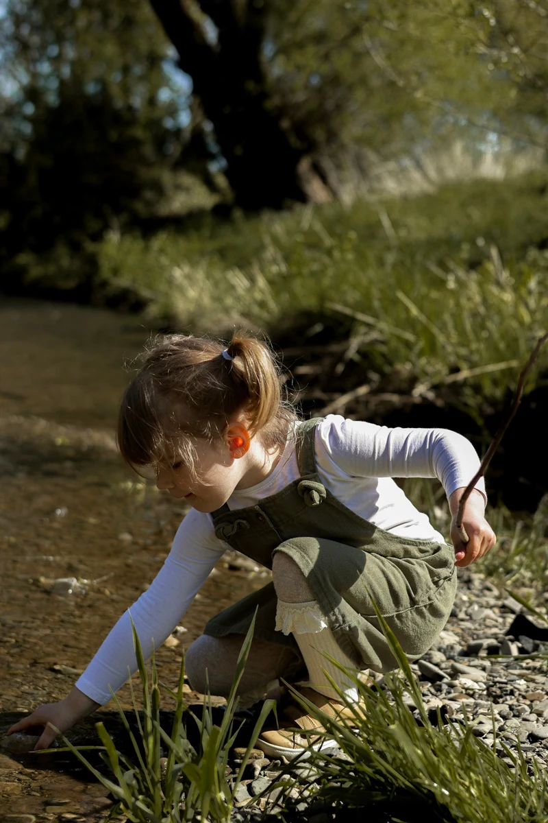 Mädchen hockt am Bachufer und greift mit der Hand ins flache Wasser, in der anderen Hand ein Stock